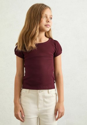 Junges Mädchen mit schulterlangem hellbraunem Haar trägt ein kurzärmeliges, dunkelrotes Shirt und cremefarbene Hosen und blickt leicht nach links.