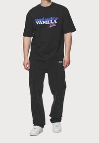 Camiseta negra con el gráfico colorido "Pecador Vanilla Super Soft!", combinada con jeans negros y zapatillas blancas. Estilo urbano simple y casual.