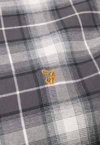 Tela a cuadros en negro, blanco y gris con un acento bordado en amarillo con la letra 'F'. La textura parece lisa con un patrón tejido.