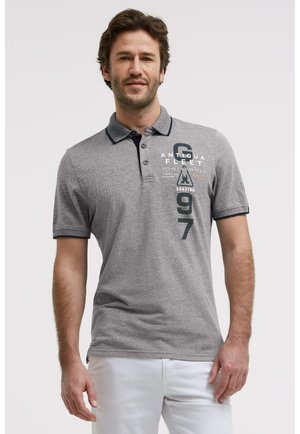 Mann trägt ein graues kurzärmliges Polo mit dem aufgedruckten Text "Antigua Fleet 1997 Gaastra" und weiße Hose, stehend vor einfachem Hintergrund.