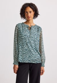 Donna con i capelli ricci indossa una blusa a maniche lunghe in teal e nero con scollo a motivo e pantaloni neri, in piedi davanti a uno sfondo neutro.