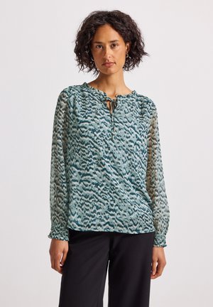Femme aux cheveux bouclés portant un blouse à manches longues noire et turquoise avec un col cravate et un pantalon noir, se tenant devant un arrière-plan uni.