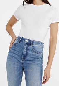 Camiseta de algodón blanca, ajustada, con mangas cortas, combinada con jeans de mezclilla azul claro de cintura alta que presentan costuras en contraste y cinco bolsillos.