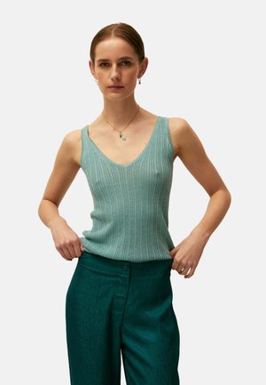 Joven mujer con el pelo peinado hacia atrás, que lleva una camiseta sin mangas de canalé azul claro y pantalones de talle alto color verde azulado oscuro, de pie con las manos a los lados.