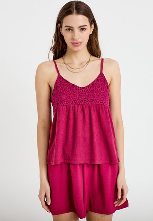 Nainen, jolla on yllään hihaton magentanvärinen pusero, jossa on virkattuja yksityiskohtia, ja siihen sopivat väljät shortsit, poseeraa yksinkertaisen vaalean taustan edessä.