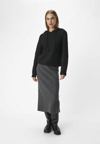 Zwarte gebreide hoodie met een textuurpatroon, gedragen met een lange grijze rok en zwarte laarzen. De outfit heeft een relaxte pasvorm en een capuchon.