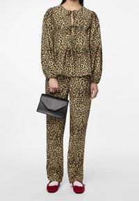 Completo in stampa leopardata composto da blusa e pantaloni realizzati in tessuto leggero; modella tiene in mano una borsa a manico superiore in pelle nera, indossando scarpe rosse slip-on.
