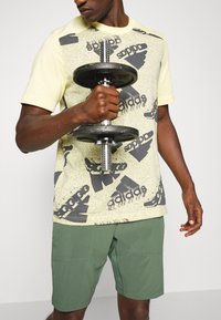 Keltainen graafinen t-paita mustalla kuviolla ja logolla, joita käytetään vihreiden shortsien kanssa. Aihe pitää mustaa käsipainoa, esitellen aktiivista vaatetustyyliä.