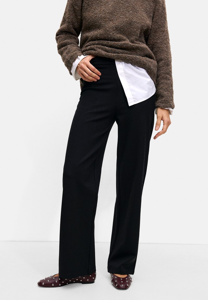 Black wide-leg trousers paired with a brown textured sweater over a white collared shirt. Burgundy studded flats complete the outfit.