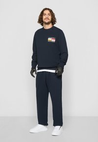 Mørkeblå hettegenser med et flerfarget logo på brystet, sammen med matchende bukser. Brukt med hvite sneakers og hansker.