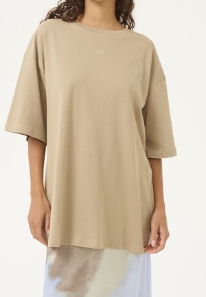 Vrouw draagt een oversized beige T-shirt met een klein logo op de borst en een lichtgekleurde rok met abstract patroon.
