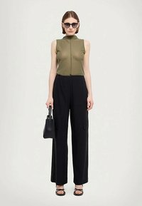 Vrouw in een mouwloze olijfgroene top, losse zwarte broek, zwarte sandalen, zonnebril en een kleine zwarte handtas, staand tegen een effen achtergrond.