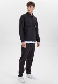 Černá bunda na zip s dvěma předními kapsami, elastickými manžetami a límcem, v kombinaci s černými kalhotami a bílými teniskami.