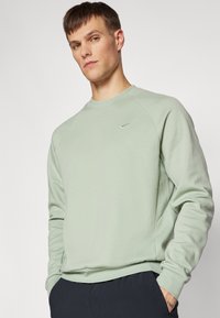 Felpa verde chiaro con collo a giro e maniche raglan, decorata con un discreto logo Nike. Tessuto morbido e vestibilità informale. Indossata con pantaloni scuri.