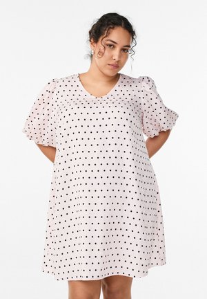 Lokkis juustega naine, kes kannab heledat roosat kleiti mustade täppidega ja padjanditega varrukatega, seisab ühevärvilise valge tausta ees.