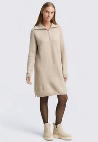 Robe pull beige tricotée avec col à zip, manches longues, fentes latérales, portée avec des bottines claires et des collants noirs.