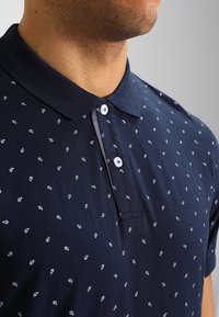 Námořnické modré polo tričko s límečkem, zdobené jemným bílým vzorem paisley. Vyrobeno z měkké tkaniny, obsahuje knoflíky pro zapínání.