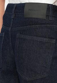 Tamsiai mėlynos džinsinės kelnės su tekstūruotu paviršiumi, turinčios juodą odinį juosmens pleistrą, ant kurio išspaustas užrašas "PEGADOR", ir standartinės galinės kišenės.