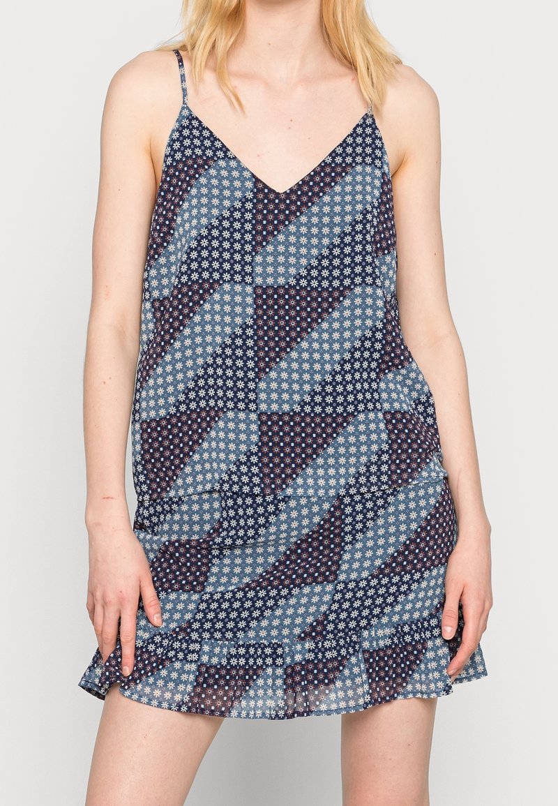 Femme portant une robe bleue sans manches avec des motifs floraux et géométriques blancs et de fines bretelles.