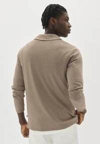Joven con trenzas cortas vestido con una camisa de manga larga beige y pantalones blancos, mirando de lado frente a un fondo blanco liso.