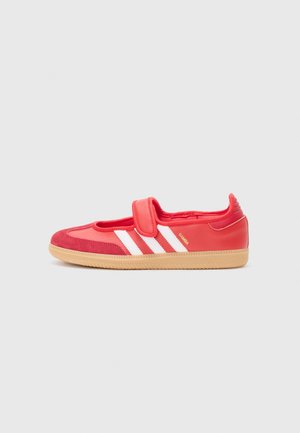 Rode Adidas Samba schoen met witte strepen, klittenbandsluiting en rubberen zool, zijaanzicht op een effen grijze achtergrond.