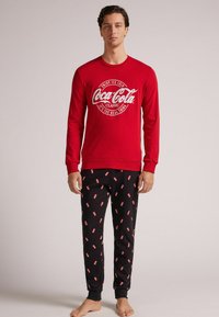 Camisa roja de manga larga con el logo de Coca-Cola, combinada con pantalones de chándal negros que presentan un patrón repetido de pequeñas latas de Coca-Cola.