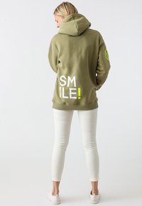 Olijf groene hoodie met een groot "SMILE!" grafiek in wit en geel op de achterkant, met een voorste kangoeroezak en een capuchon met trekkoord.