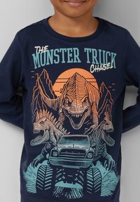 Marineblauw shirt met lange mouwen, dat een grafisch ontwerp van twee dinosaurussen en een monstertruck toont, met oranje en teal accenten tegen een bergachtergrond.