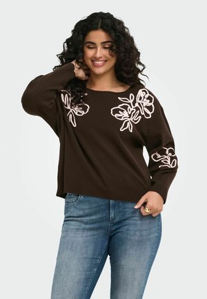 Kvinde med krøllet mørkt hår, smilende, iført en mørkebrun sweater med hvid blomsterbroderi og blå jeans, poserer med den ene hånd bag hovedet.