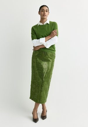 Woman standing with arms crossed wearing a green sweater, white shirt, shiny green textured skirt, and black pointed heels against a plain background.