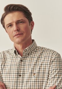 Cămașă cu nasturi, în carouri maro și bej, având un mic logo brodat cu un urs pe piept. Cu guler și mâneci scurte.