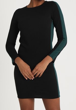 Sort tætsiddende kjole med lange ærmer og mørkegrønne striber langs ærmer og sider, båret af en person med hænderne foldet foran.