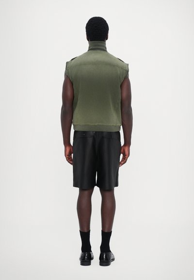 Veste sans manches vert olive, ourlet côtelé, portée sur un short en cuir noir, associée à des chaussettes et des chaussures noires, vue de l'arrière.