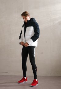 Polstret jakke med sorte og hvide sektioner, parret med sorte leggings med et logo og røde træningssko med hvide accenter.