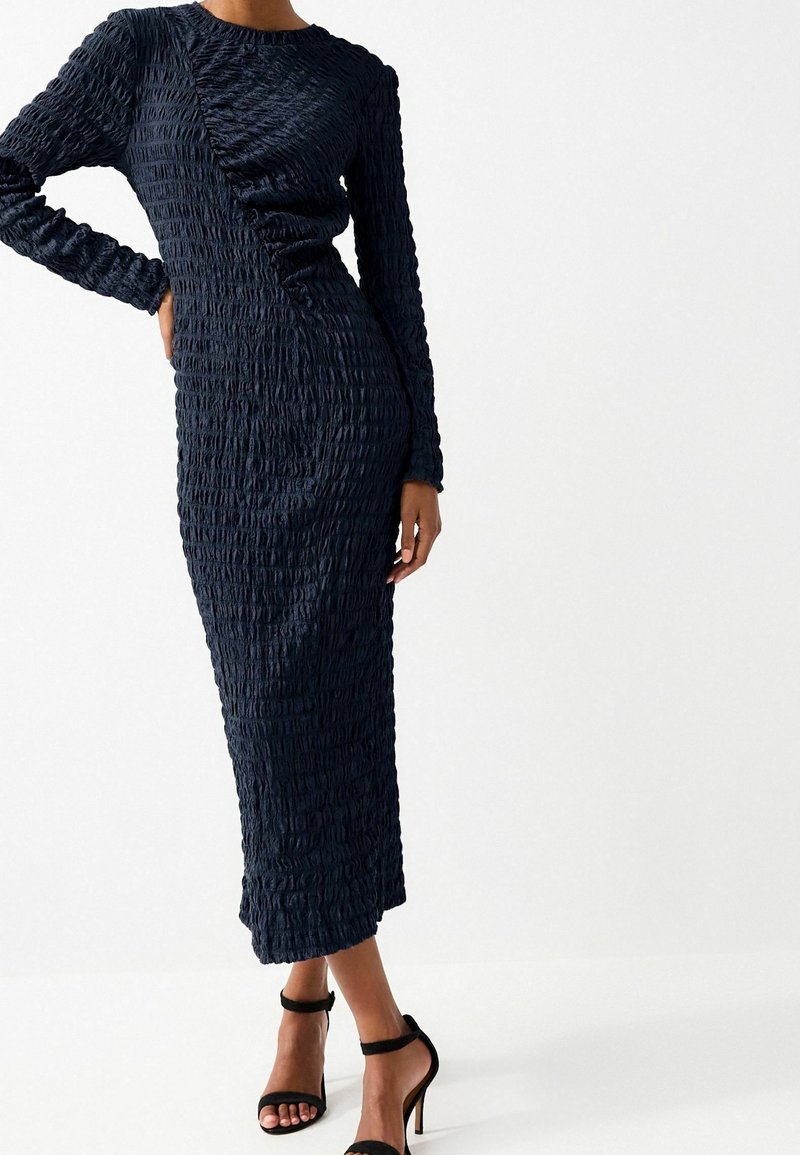 Femme portant une robe midi texturée bleu foncé à manches longues avec des détails froncés, associée à des sandales noires à talons hauts sur fond blanc.