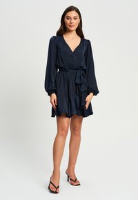 Mornarska obleka z zavojem in dolgimi rokavi, s pasom na vezanje in plastičnim robom. Narejena je iz gladkega materiala z rahlo sijočim finišem.