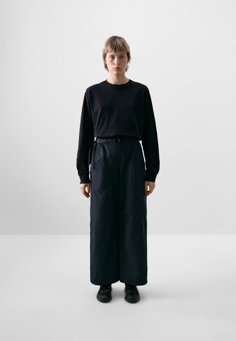 Zwart langemouwen shirt gecombineerd met brede, hoog-getailleerde zwarte broeken. Broeken hebben riemlussen en twee zijzakken.