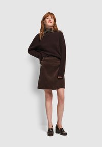 Kobieta nosząca ciemnobrązowy, oversize'owy sweter, paskowany golf, brązową spódnicę z kordurowej tkaniny z guzikami oraz ciemnobrązowe skórzane mokasyny na obcasie, stojąca.