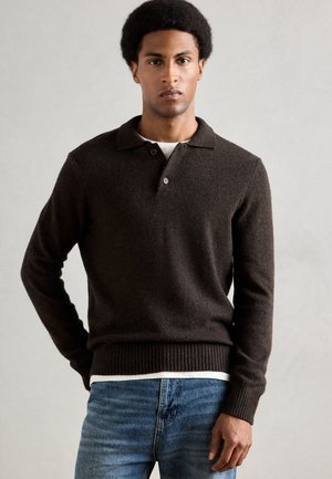 Brun strikket pullover med krave, to-knap lukning og ribbede manchetter. Båret over en hvid t-shirt, kombineret med blå jeans.