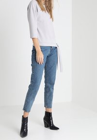 Lichtgrijze top met driekwartmouwen en een gestrikte taille, gecombineerd met blauw, nauwsluitende jeans en zwarte, hoge laarzen met hakken. Gladde textuur.