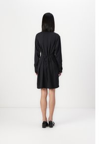 Robe noire à manches longues avec ceinture à nouer. Présente un motif texturé, un ourlet légèrement arrondi, et tombe au-dessus du genou. Portée avec des chaussures noires.