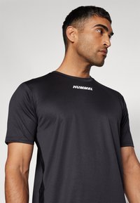 Musthave mustar t-särk, mis on valmistatud kergest kangast, lühikeste varrukatega ja ümmarguse kaelusega. Rinnal on valge "HUMMEL" logo.