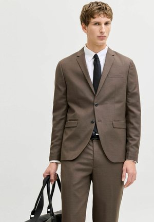 Jonge man in een bruin pak, wit overhemd, zwarte stropdas, met een zwarte leren tas tegen een effen lichte achtergrond.