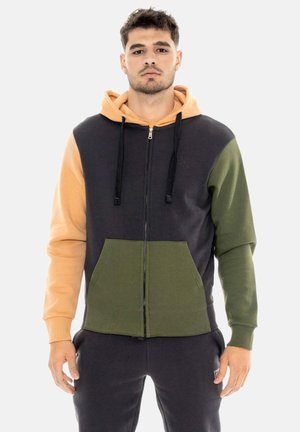 Jonge man die een color-block zip-up hoodie draagt met beige, zwarte en olijfgroene delen, staand tegen een effen achtergrond.