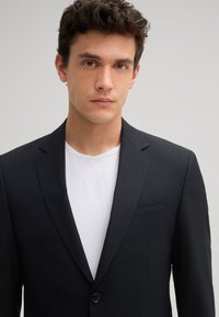 Zwarte op maat gemaakte blazer met een enkele knoop, notch revers en een borstzak, gedragen over een witte ronde hals t-shirt. Glad oppervlak.