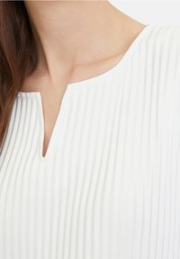 Top bianco plissettato con scollatura a V. La trama del tessuto è liscia e presenta righe verticali, enfatizzando un design leggero e arioso.