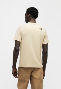 Μπεζ βαμβακερό T-shirt με κοντά μανίκια, στρογγυλή λαιμόκοψη και μικρή μαύρη λεπτομέρεια λογότυπου στο πίσω μέρος, σε συνδυασμό με καφέ παντελόνι καρό.