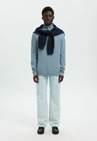 Muškarac u svijetloplavom dukserici sa zatvaračem, tamnoplavoj dukserici prebačenoj preko ramena, svijetloplavim trapericama i crnim cipelama, stoji nasuprot bijeloj pozadini.