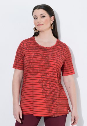 Frau mit langen dunklen Haaren, die große Creolen und ein rotes kurzärmeliges Shirt mit horizontalen Streifen und Blumenmuster trägt.
