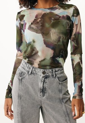 Femme portant un haut à manches longues à motif aquarelle dans des tons verts et bruns avec un jean taille haute gris clair.